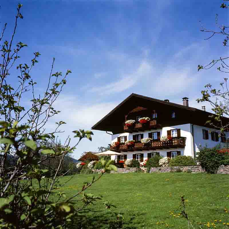 Ein traditionelles oberbayerisches Hotelgebäude mit weißer Fassade, dunklen Holzbalkonen und üppigem Blumenschmuck steht auf einer grünen Wiese unter strahlend blauem Himmel.