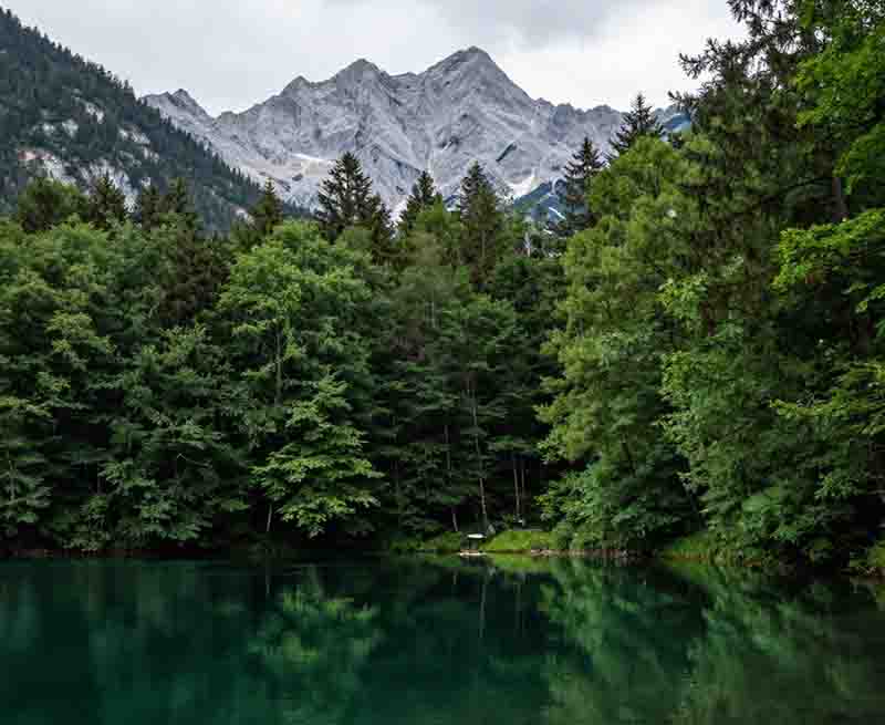 Garmisch Partenkirchen Landschaft mit einem dunkelgrünen See im Vordergrund, der die umliegenden bewaldeten Ufer und die grauen Berggipfel im Hintergrund widerspiegelt.