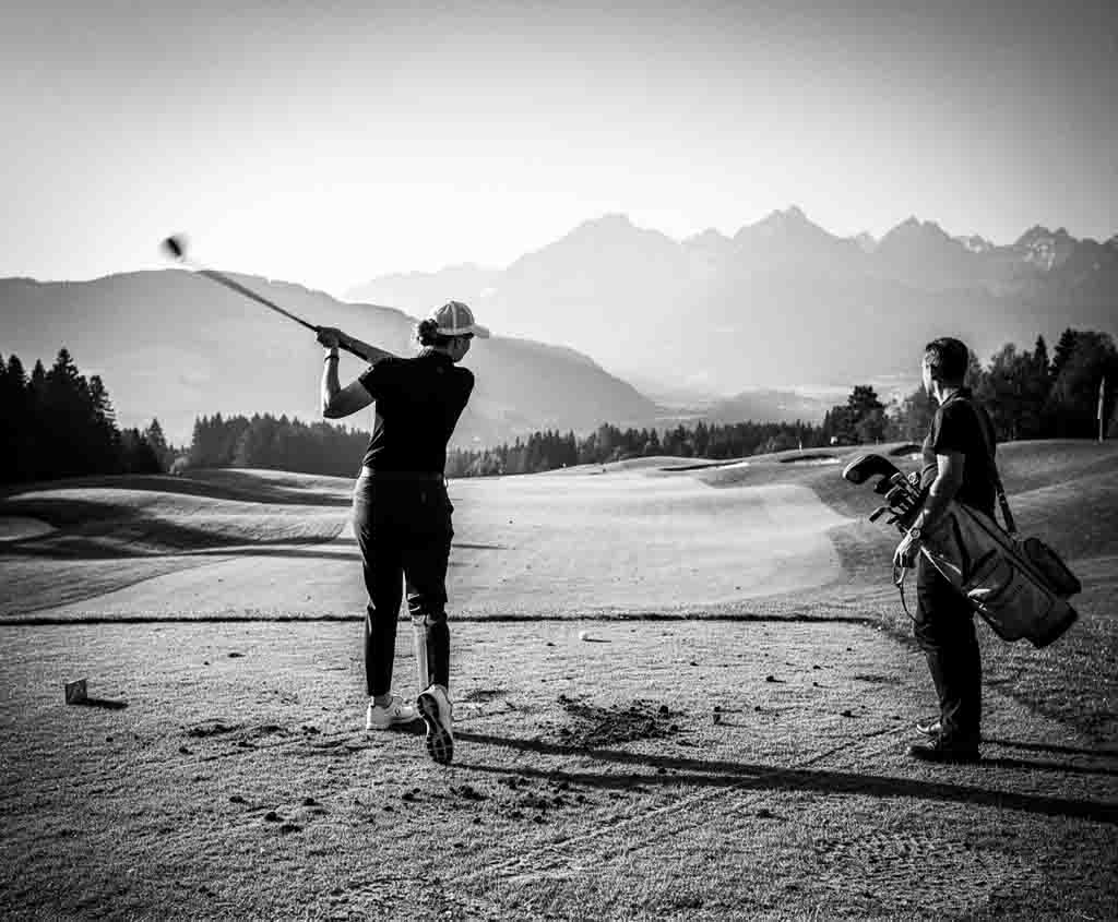Schwarz-Weiß-Fotografie einer Golferin beim dynamischen Abschlag auf einem Tee in Oberbayern, während ein Caddie mit Golftasche rechts im Bild zuschaut. Im Hintergrund erstreckt sich ein weitläufiges Fairway vor einer imposanten Gebirgskette der Bayerischen Alpen im Gegenlicht.