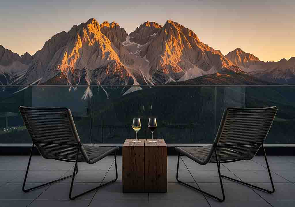 Zwei moderne Design-Sessel auf einer Hotelterrasse in Oberbayern mit Glasbalustrade blicken auf ein imposantes Gebirgspanorama bei Sonnenuntergang. Auf einem rustikalen Holzbeistelltisch stehen ein Glas Weißwein und ein Glas Rotwein.