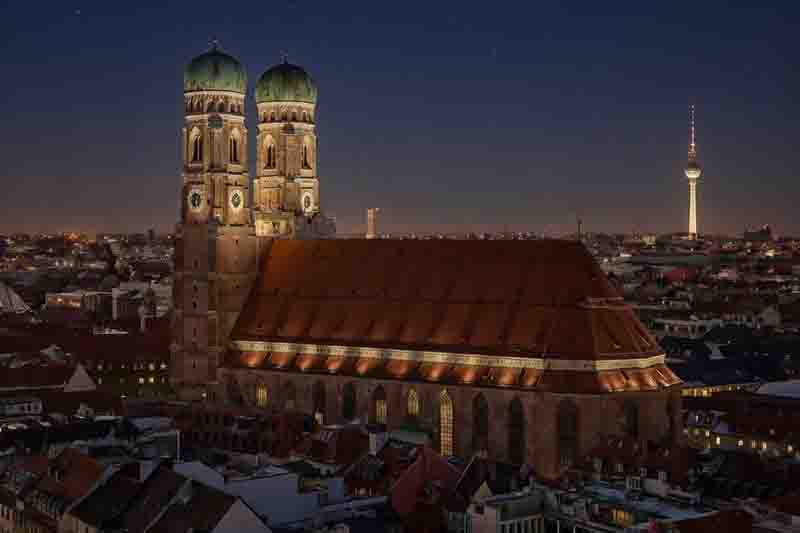 Nächtliche Stadtansicht, die die Münchener Frauenkirche mit ihren markanten Zwiebeltürmen im Vordergrund und den Berliner Fernsehturm im beleuchteten Hintergrund kombiniert.