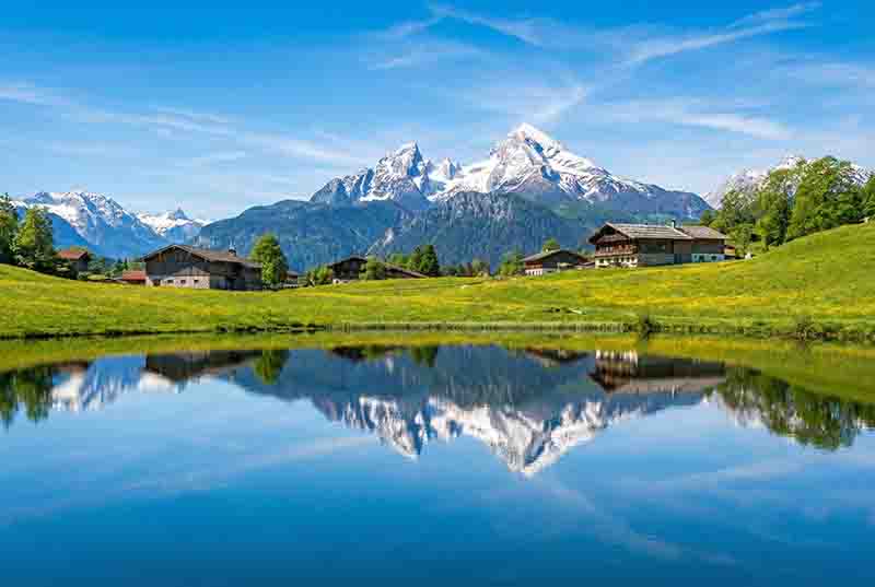 Ein idyllisches Oberbayrisches Alpenpanorama unter strahlend blauem Himmel. Im Vordergrund erstreckt sich ein stiller, tiefblauer See, der die gesamte Szenerie perfekt spiegelt.