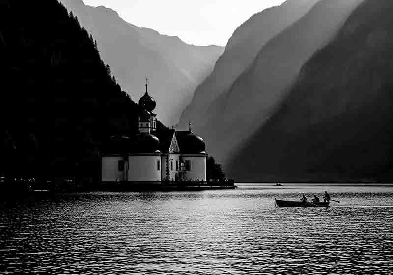 Dies ist ein dramatisches Schwarz-Weiß-Foto der Kapelle St. Bartholomä am Königssee. Die Kapelle mit ihren markanten Zwiebeltürmen steht hell beleuchtet am Ufer vor einer gewaltigen, dunklen Bergkulisse.